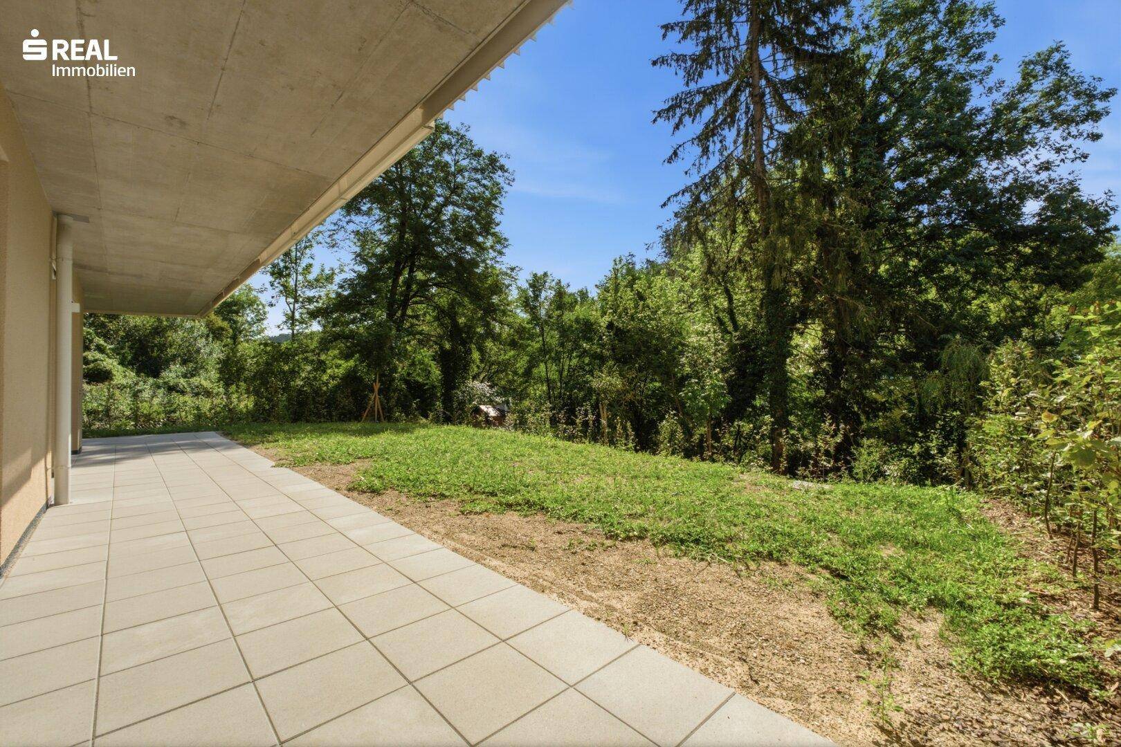 Grosse gepflasterte Terrasse neben einer weitläufigen Grünfläche, umgeben von Bäumen und Natur.