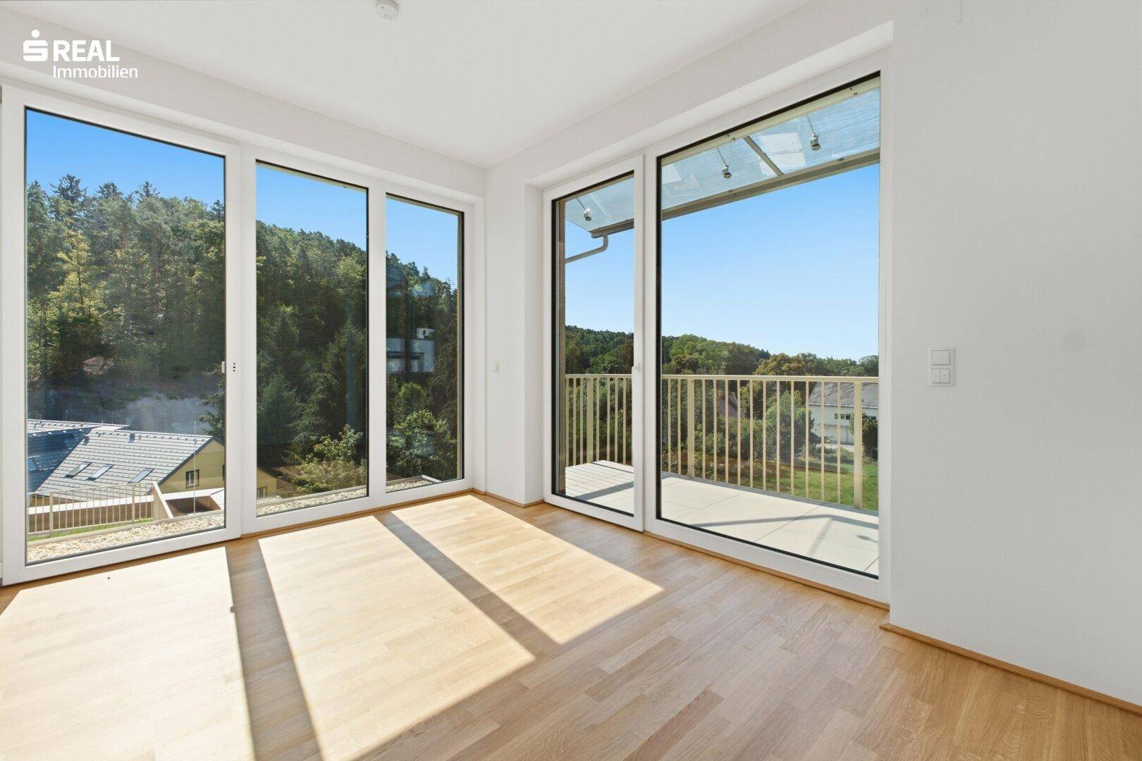 Heller Raum mit Holzfußboden, großen Fenstern und Zugang zu einem Balkon.