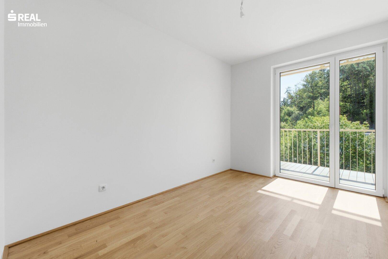 Leeres Schlafzimmer mit Holzboden und großem Fenster, das viel natürliches Licht hereinlässt.
