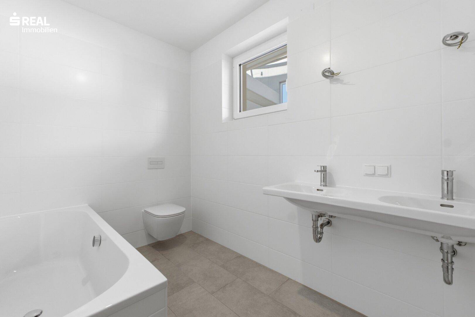 Modernes Badezimmer mit weißen Fliesen, Doppelwaschbecken, Badewanne und Fenster für Tageslicht.