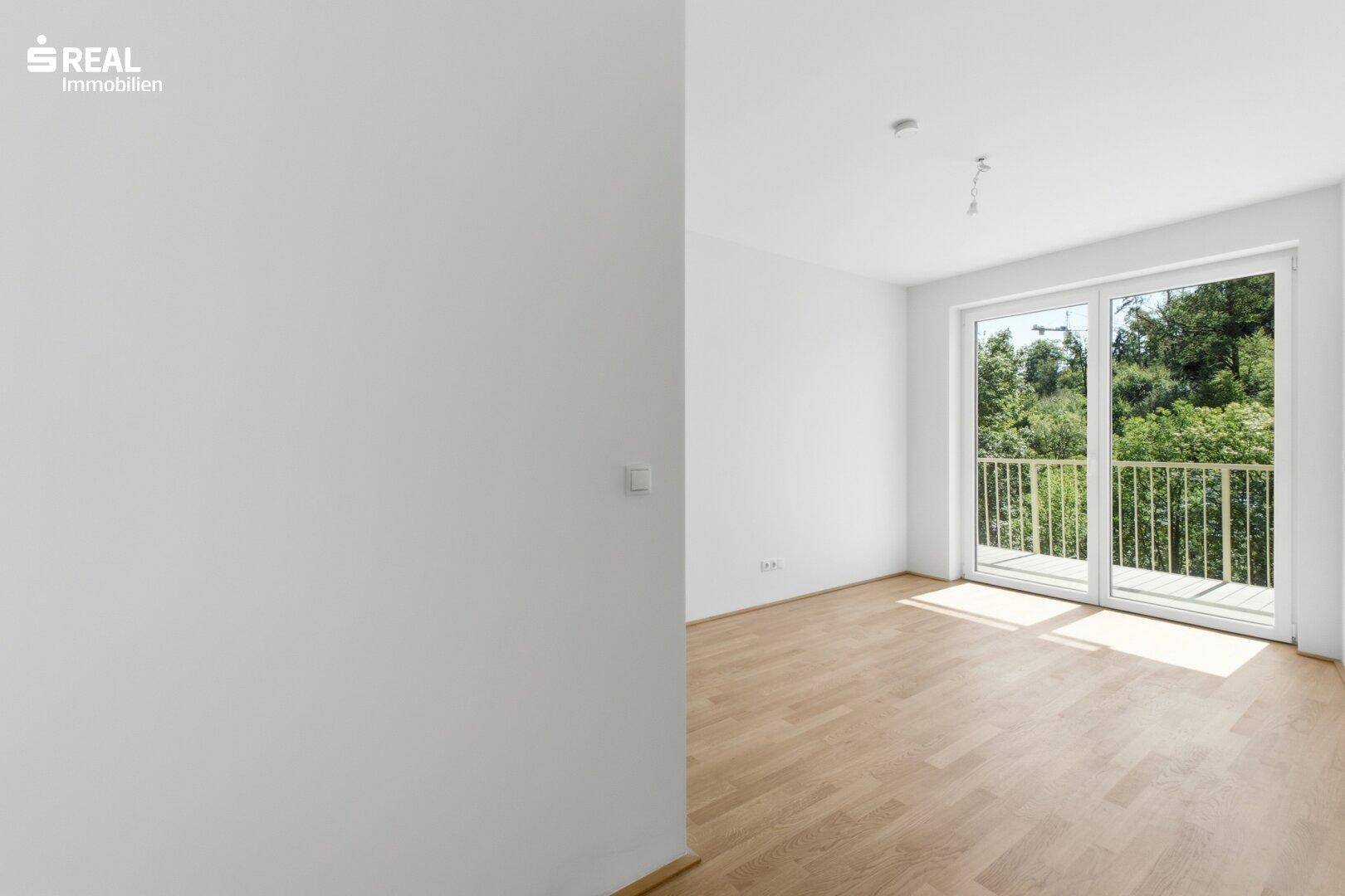 Lichtdurchflutetes Zimmer mit Holzboden und Balkonzugang, bietet einen schönen Blick ins Grüne.