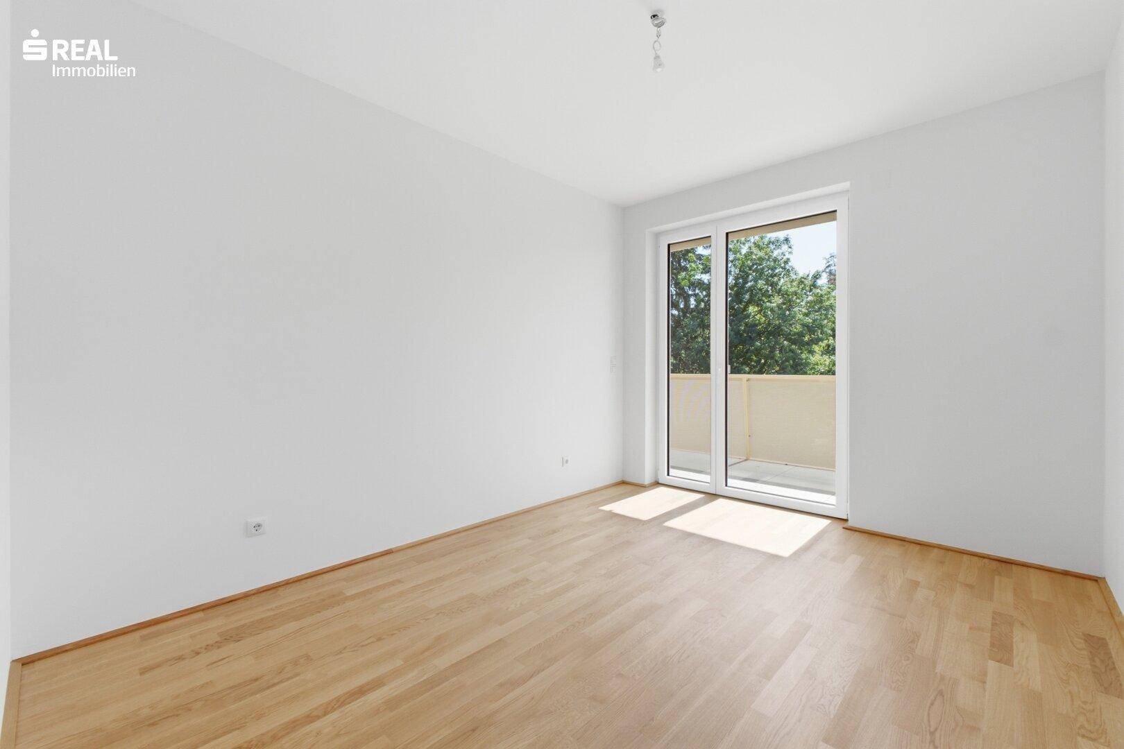 Unmöbliertes Zimmer mit hellem Parkettboden und Zugang zu einem Balkon, ideal als Schlafzimmer.