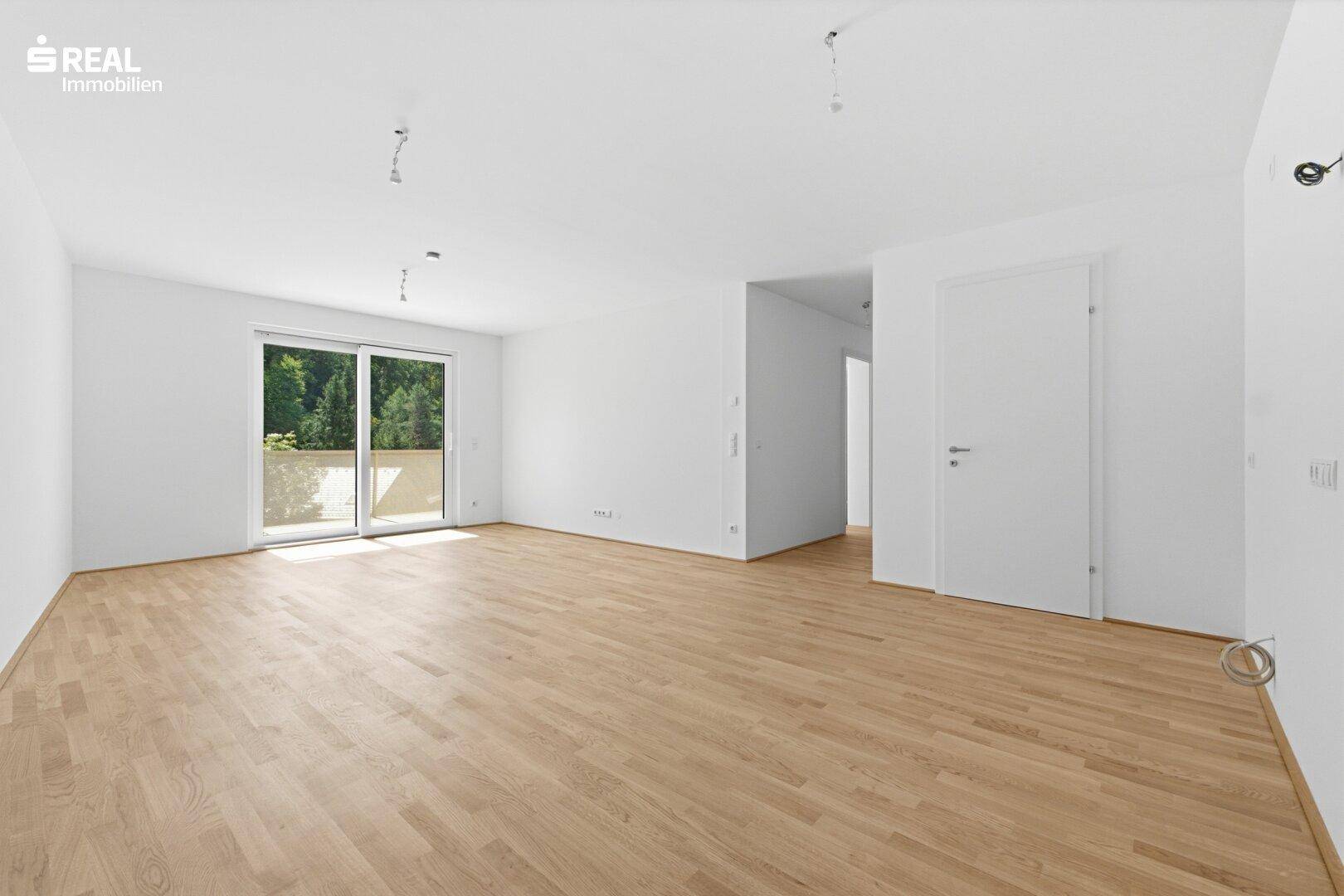 Helles, leeres Wohnzimmer mit Parkettboden und Zugang zu einem Balkon mit viel Tageslicht.