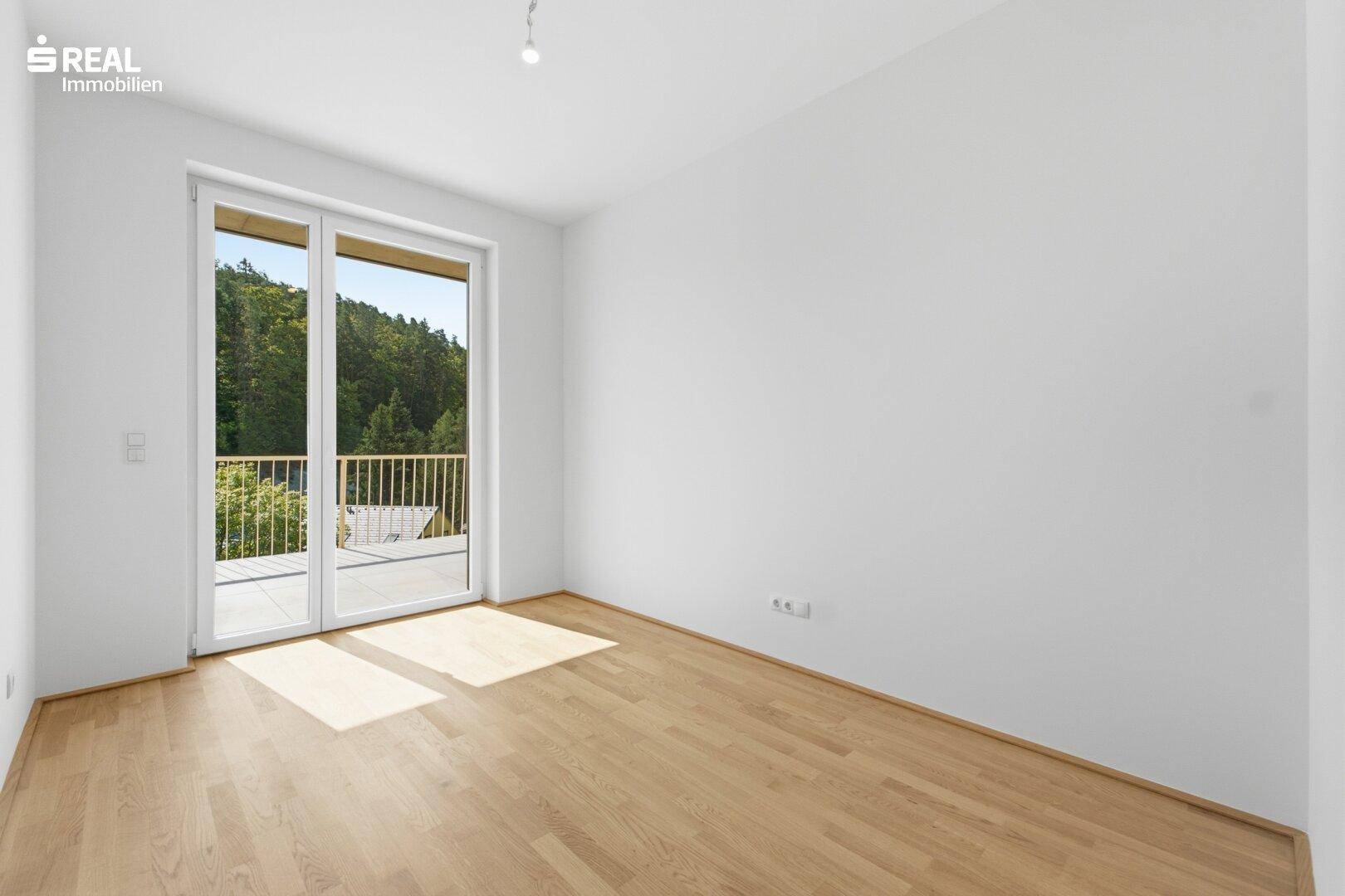 Lichtdurchflutetes Zimmer mit Holzboden und Zugang zu einem Balkon mit grüner Aussicht.
