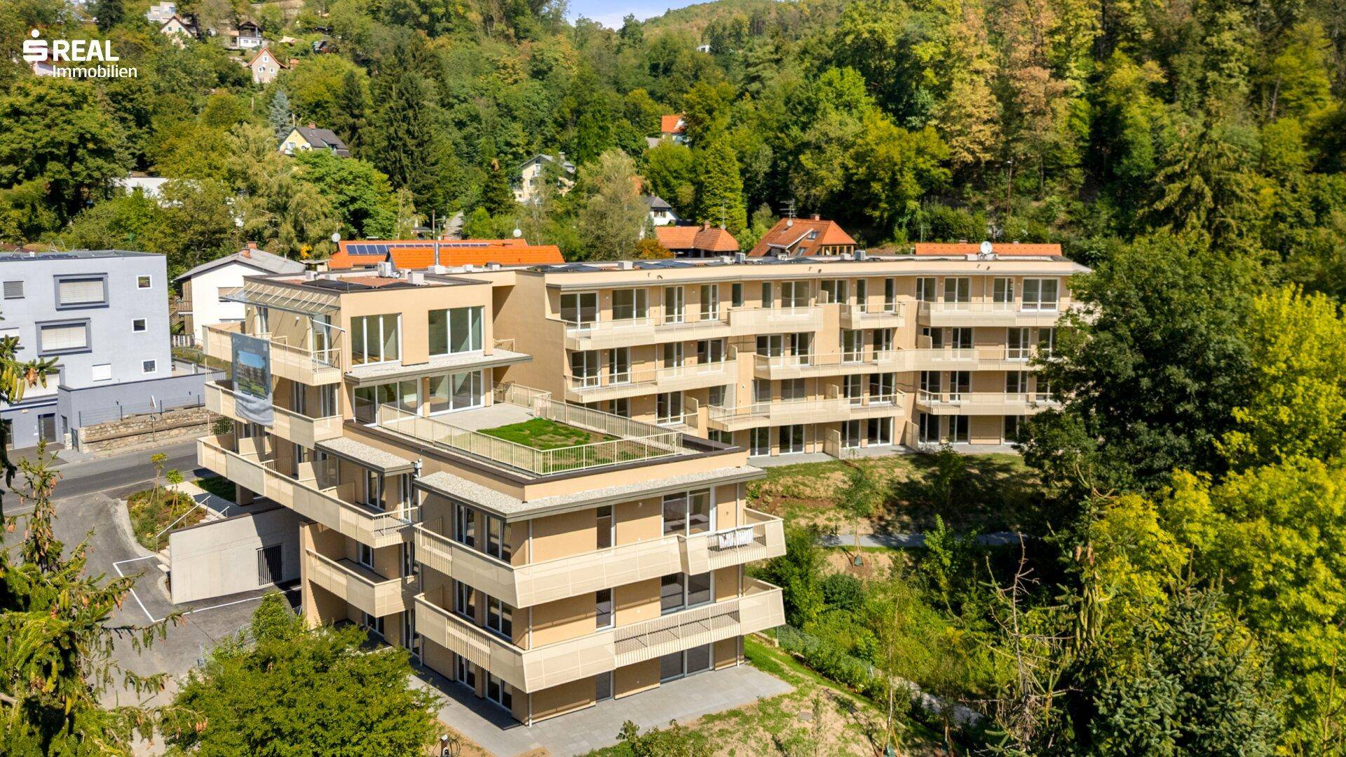 Weitläufiges Apartmentgebäude in moderner Bauweise, eingebettet in eine grüne Hügellandschaft mit vielen Bäumen.