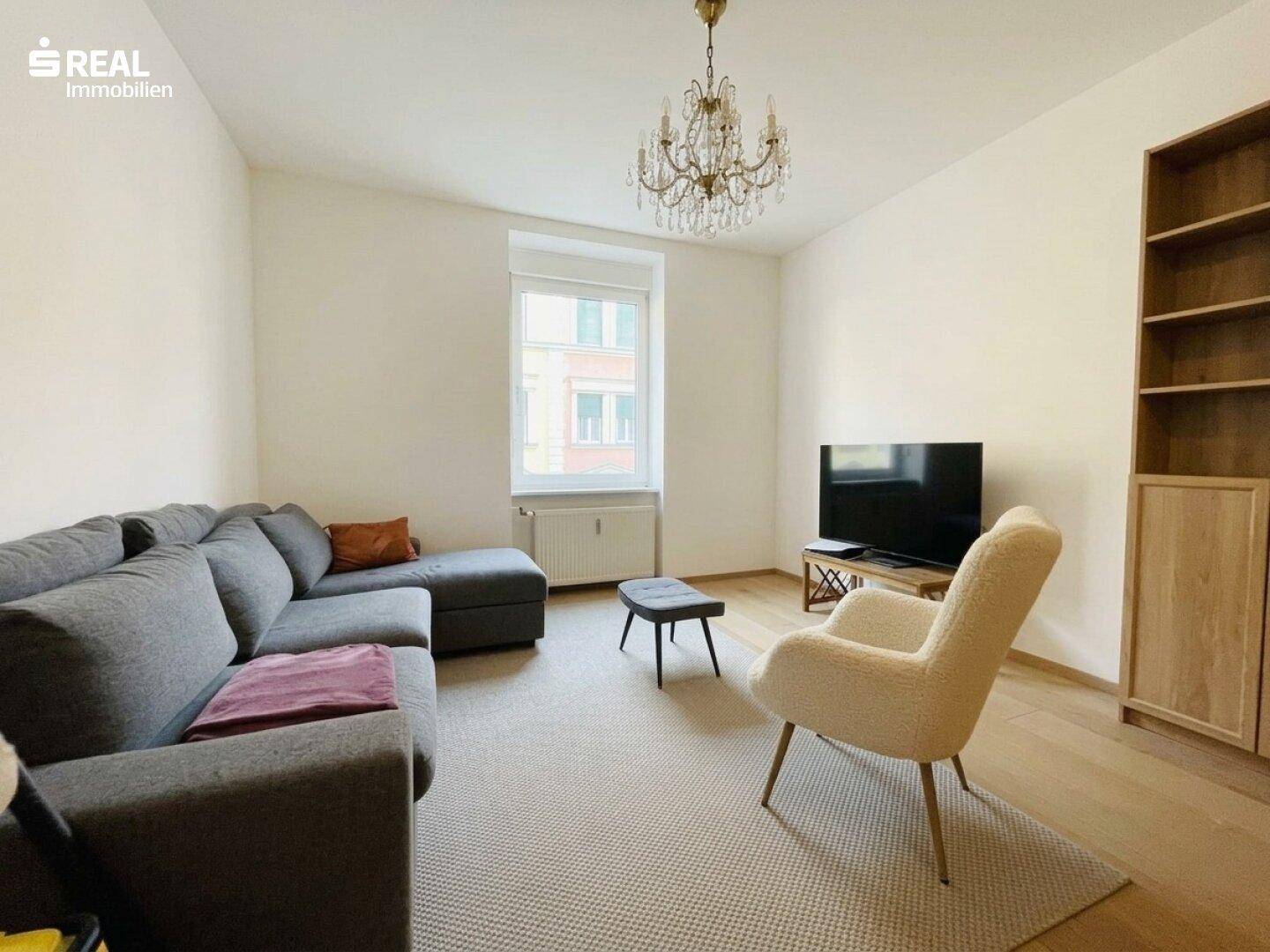 Gemütliches Wohnzimmer mit grauem Sofa, hellem Sessel, Kronleuchter und Holzboden.