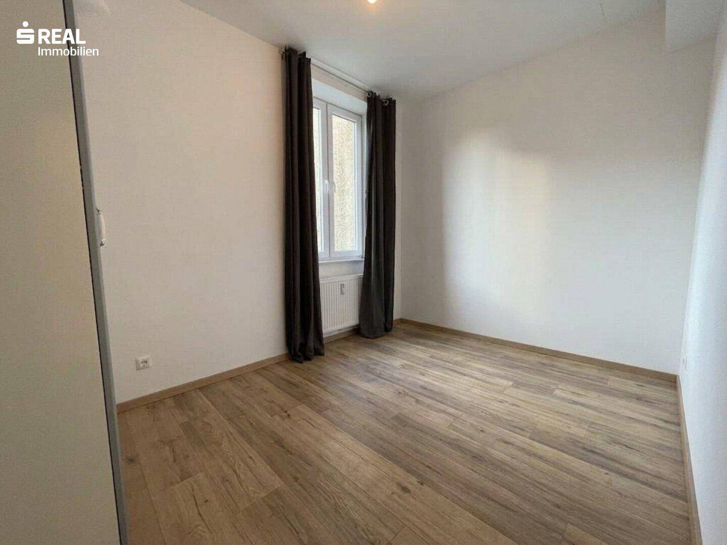 Leeres Schlafzimmer mit hellem Holzboden, weißen Wänden und dunklen Vorhängen am Fenster.