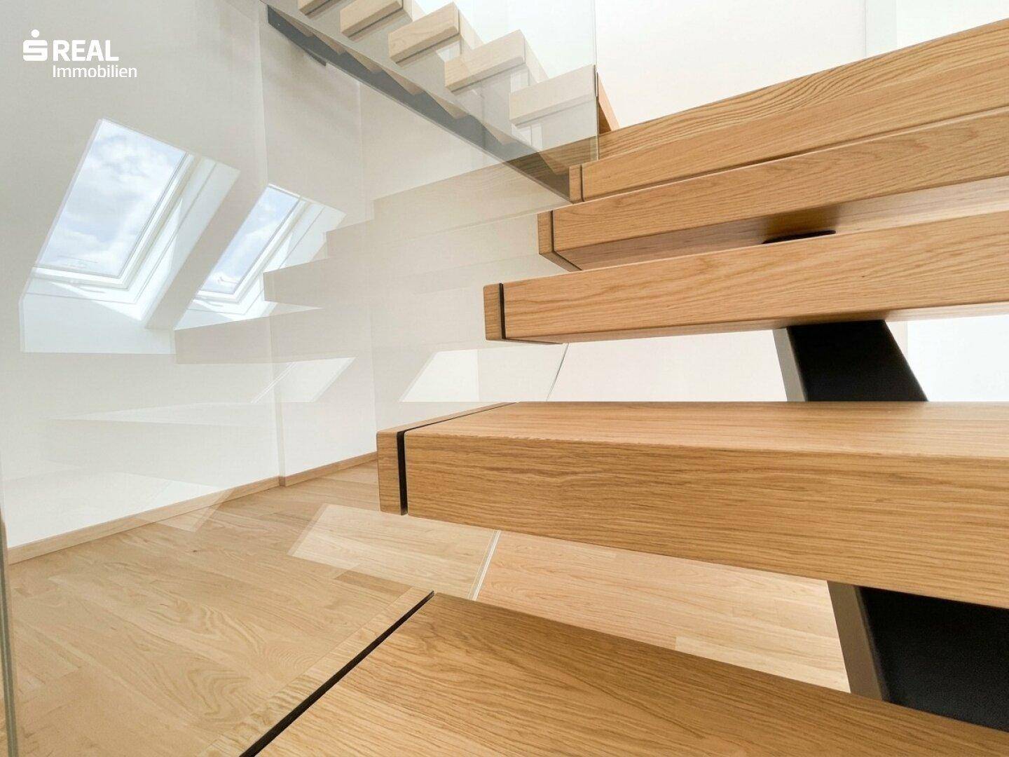 Detailaufnahme einer modernen Holztreppe mit Glasgeländer und hellen Dachfenstern im Hintergrund.