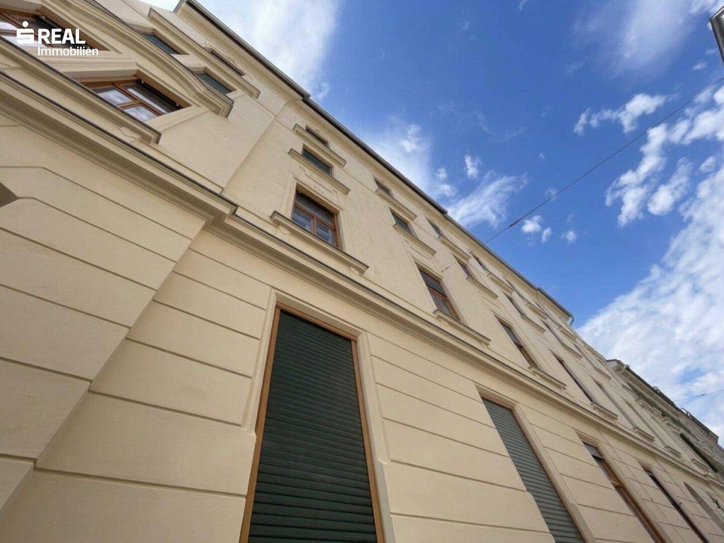 Historische Fassade eines Gebäudes mit vielen Fenstern und Rollläden unter blauem Himmel.