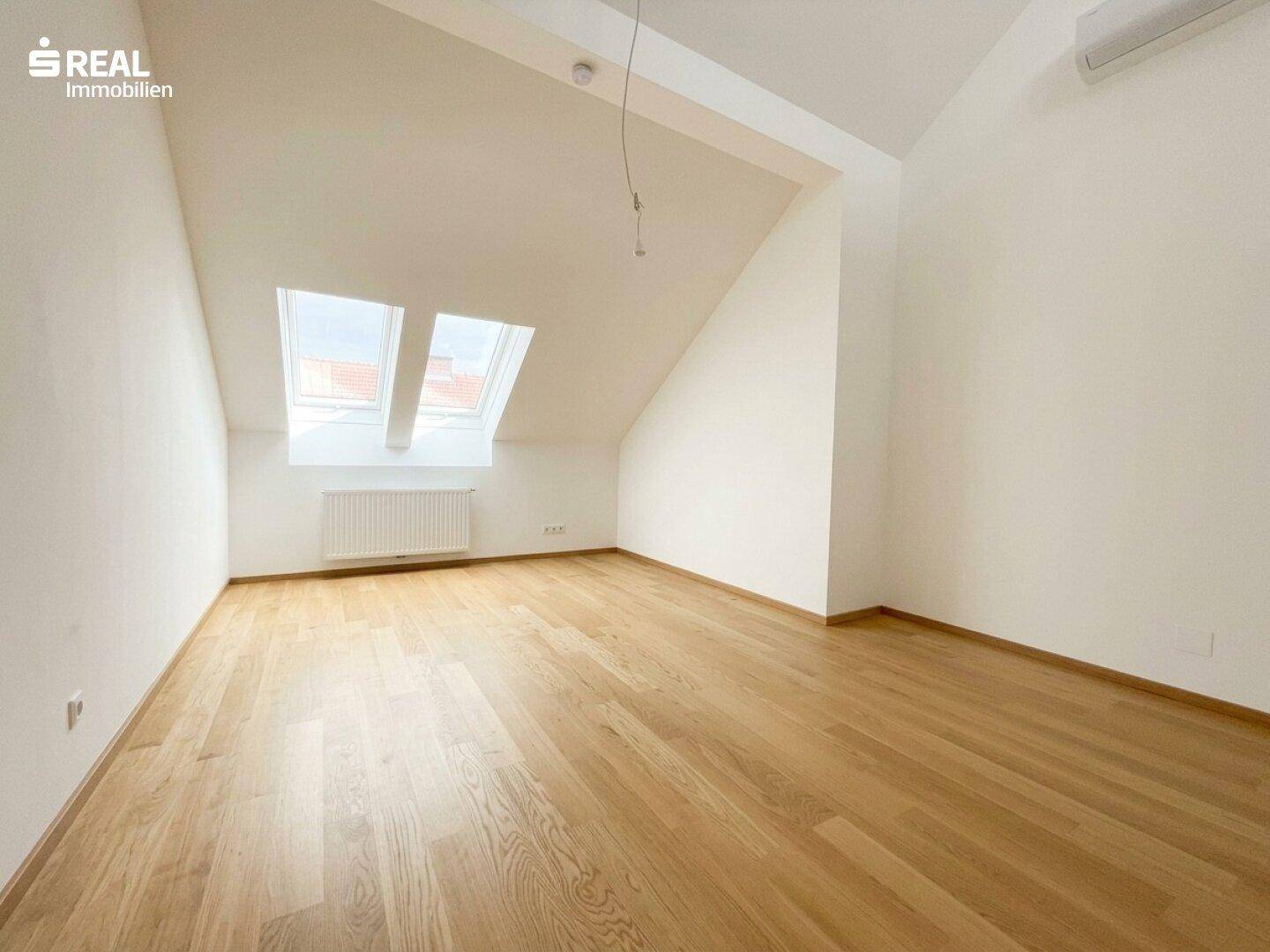 Helles Dachzimmer mit Holzboden, schrägen Wänden und zwei Dachfenstern für viel Tageslicht.