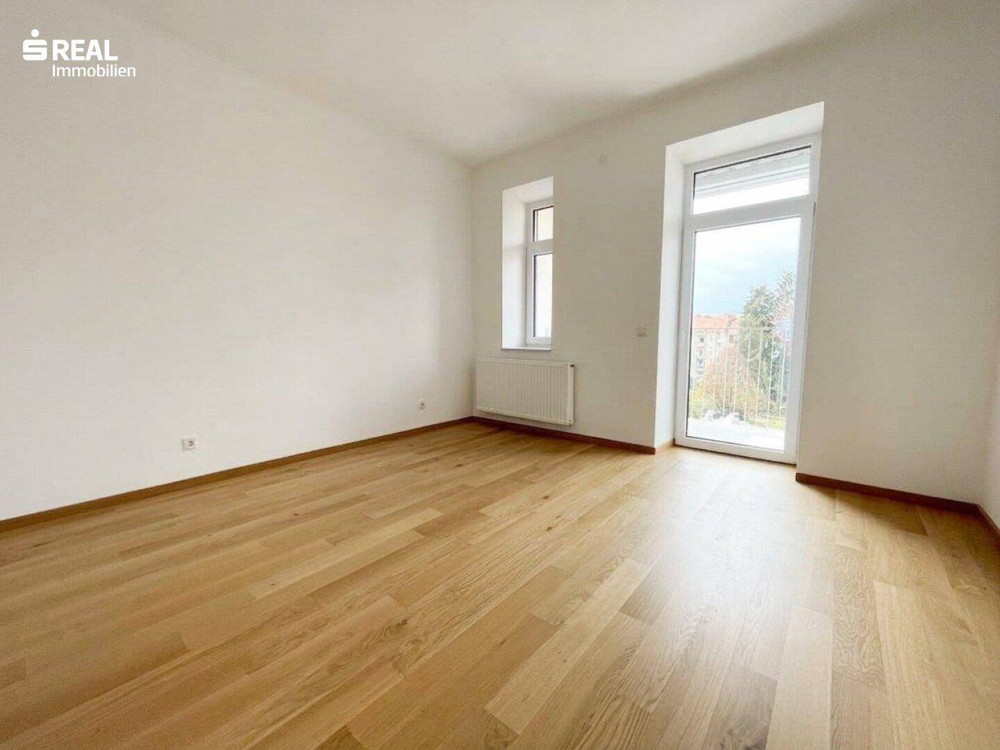 Leeres Zimmer mit Holzboden, einem Fenster und einer Tür, die zu einem französischen Balkon führt.