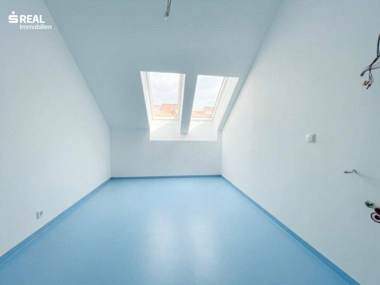 Dachzimmer mit blauer Bodenfläche, schrägen Wänden und zwei Dachfenstern für helles Tageslicht.