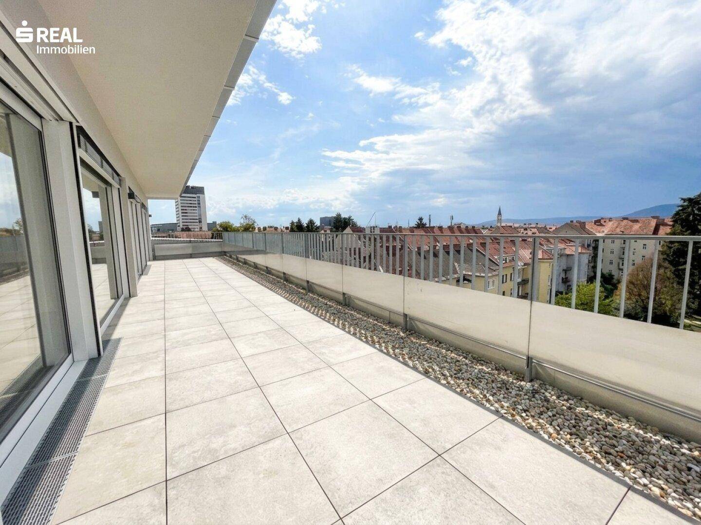 Große Dachterrasse mit Fliesenboden, Glasgeländer und weitem Blick über die Stadt.