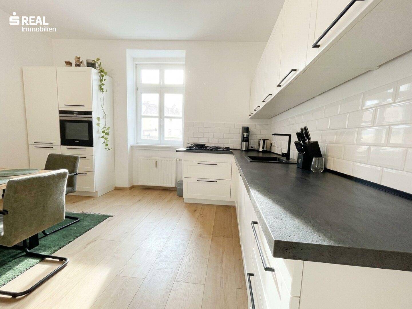 Moderne Küche mit weißer Schrankfront, dunkler Arbeitsplatte und Gasherd am Fenster.