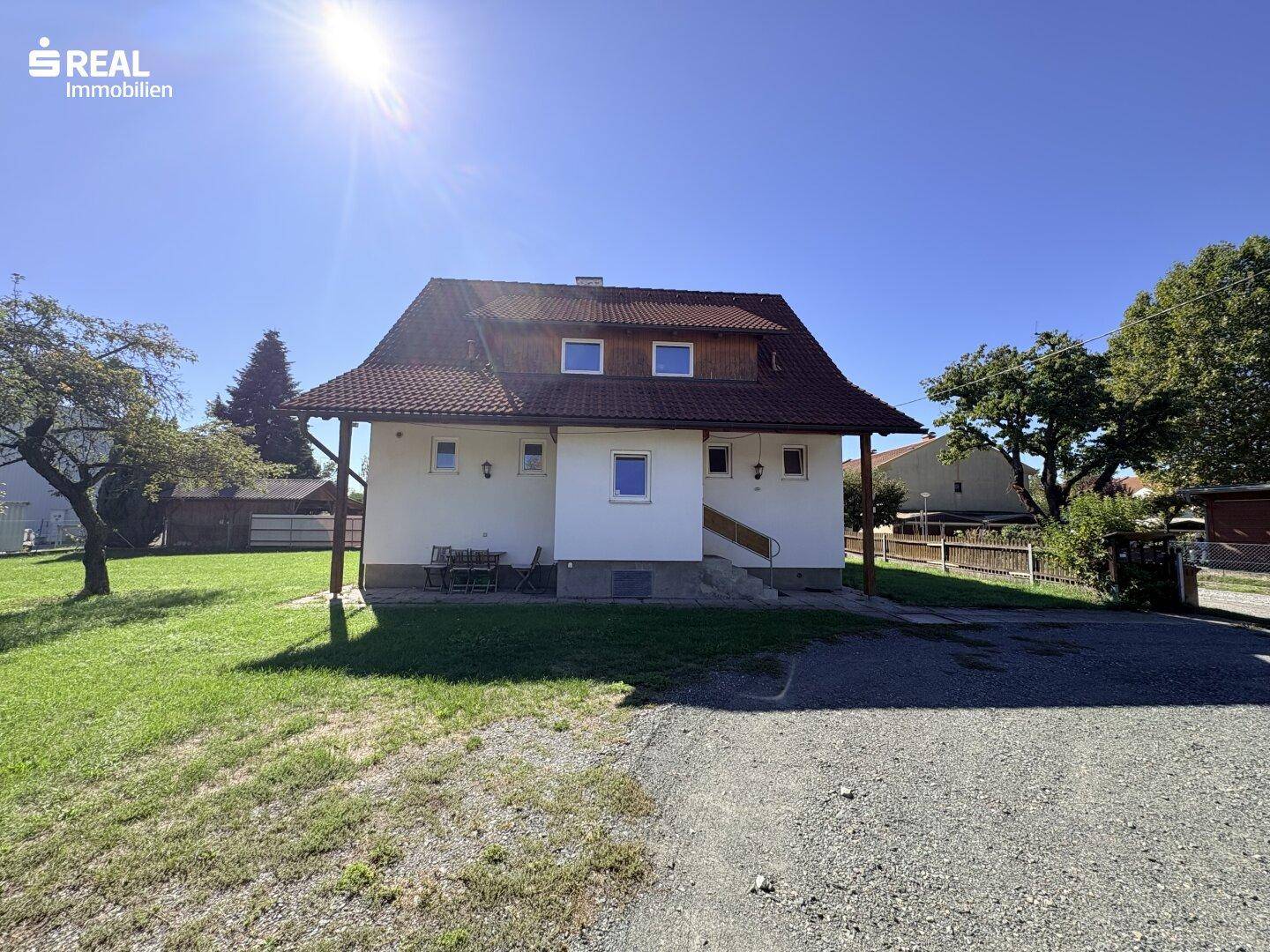 Rückansicht des Hauses mit überdachtem Sitzbereich, Kiesauffahrt und sonniger Umgebung.