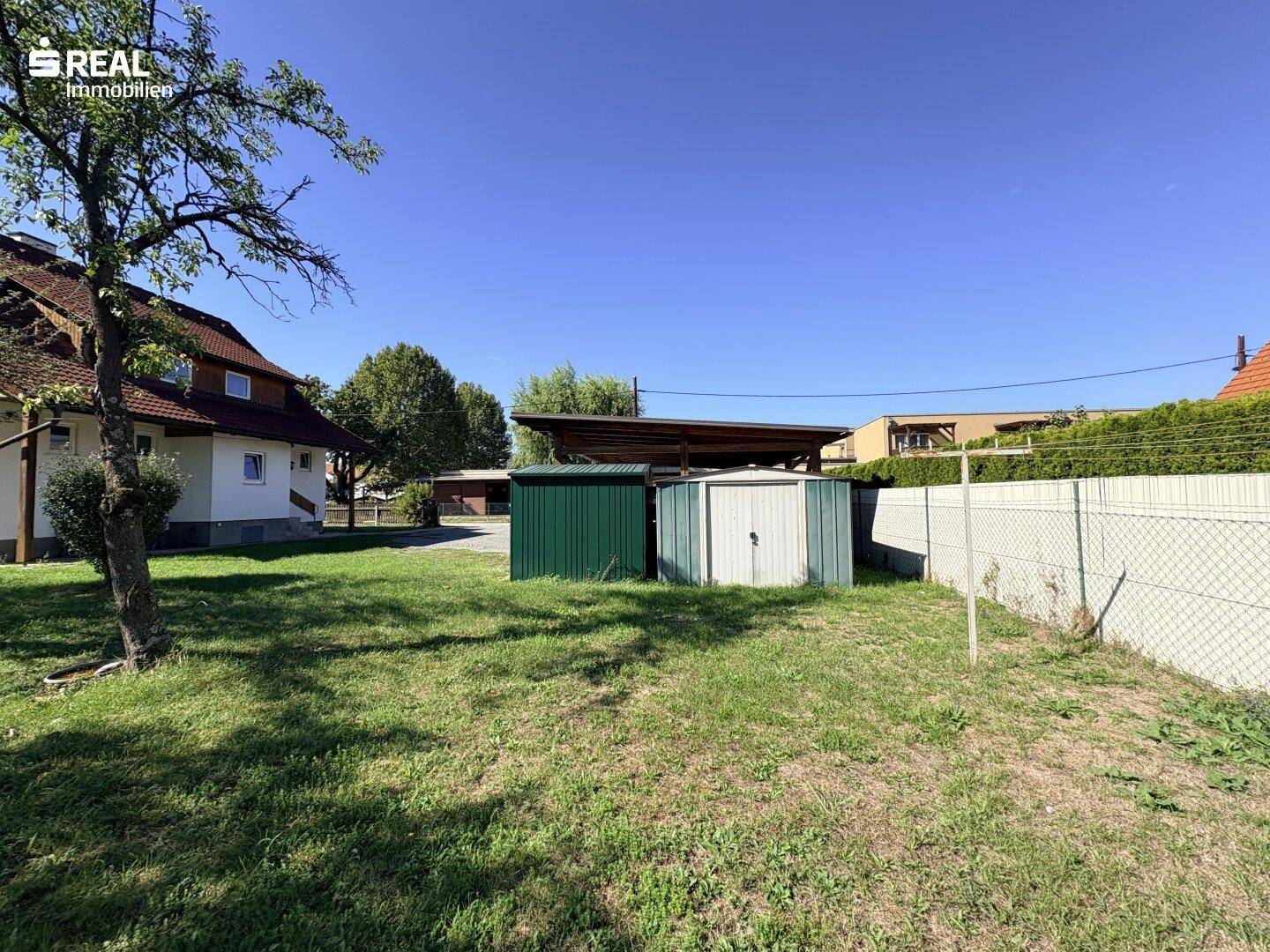 Gartenbereich mit Rasen, Gartenhäusern und einem Carport im Hintergrund des Grundstücks.