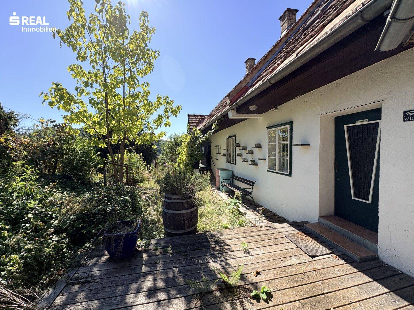 Rustikale Holzterrasse entlang der Hausfassade, umgeben von Pflanzen und einem Weinfass.