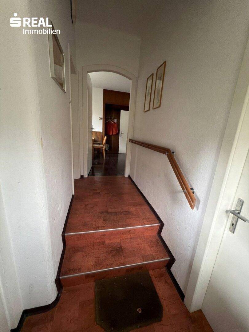Schmaler Flur mit Holztreppe und Handlauf, der zu weiteren Räumen führt.