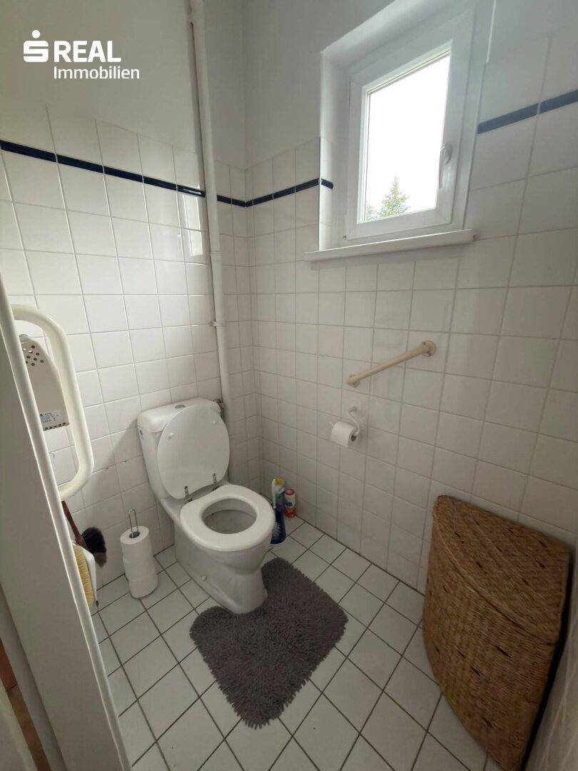 Kleines WC mit weißen Fliesen, Fenster und Haltegriffen für Barrierefreiheit, in einem funktionalen Design.
