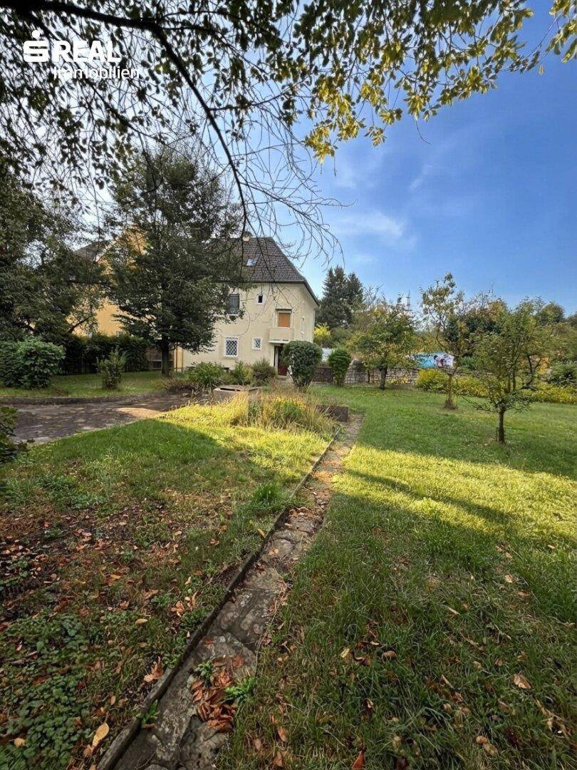 Weitläufiger Gartenbereich mit Rasenfläche und Bäumen, das Haus im Hintergrund unter blauem Himmel.