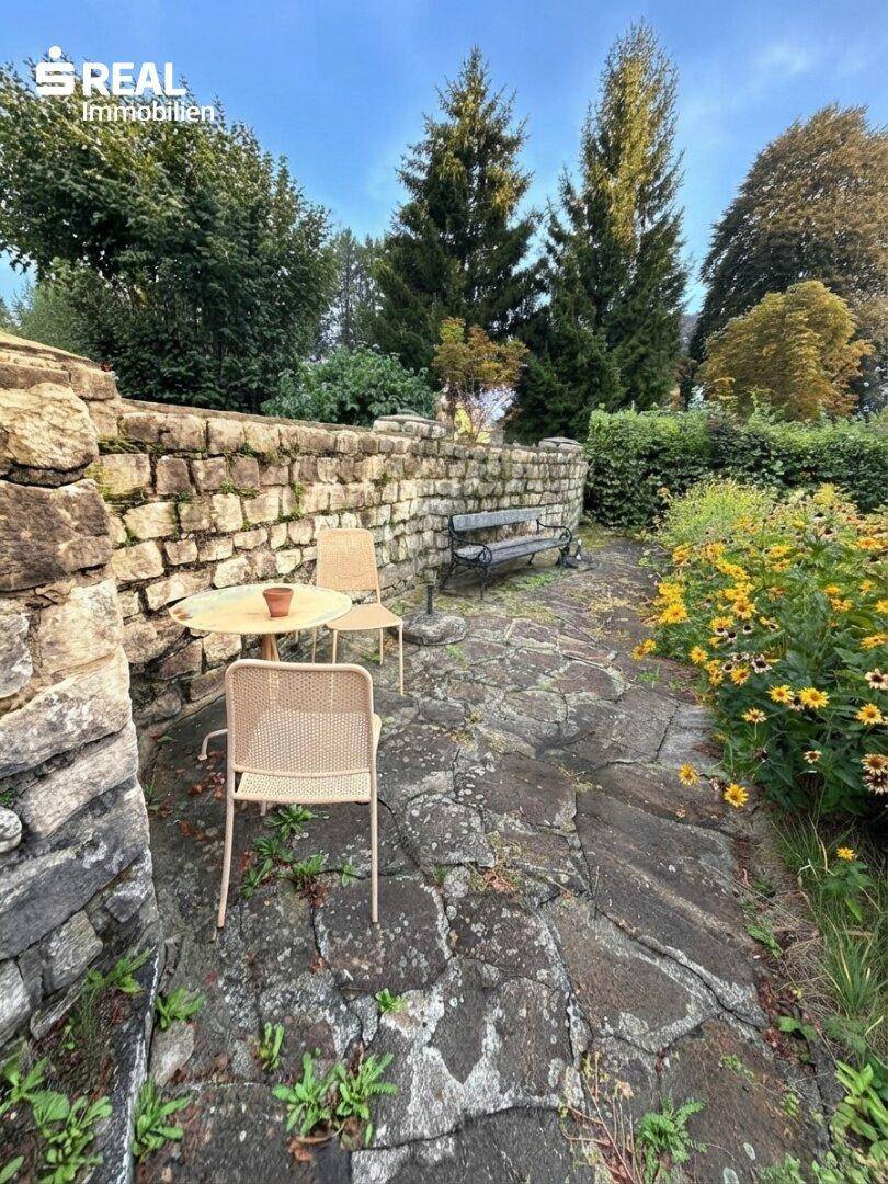 Gemütliche Steinterrasse mit Tisch und Stühlen, umgeben von Blumen und einer Steinmauer.