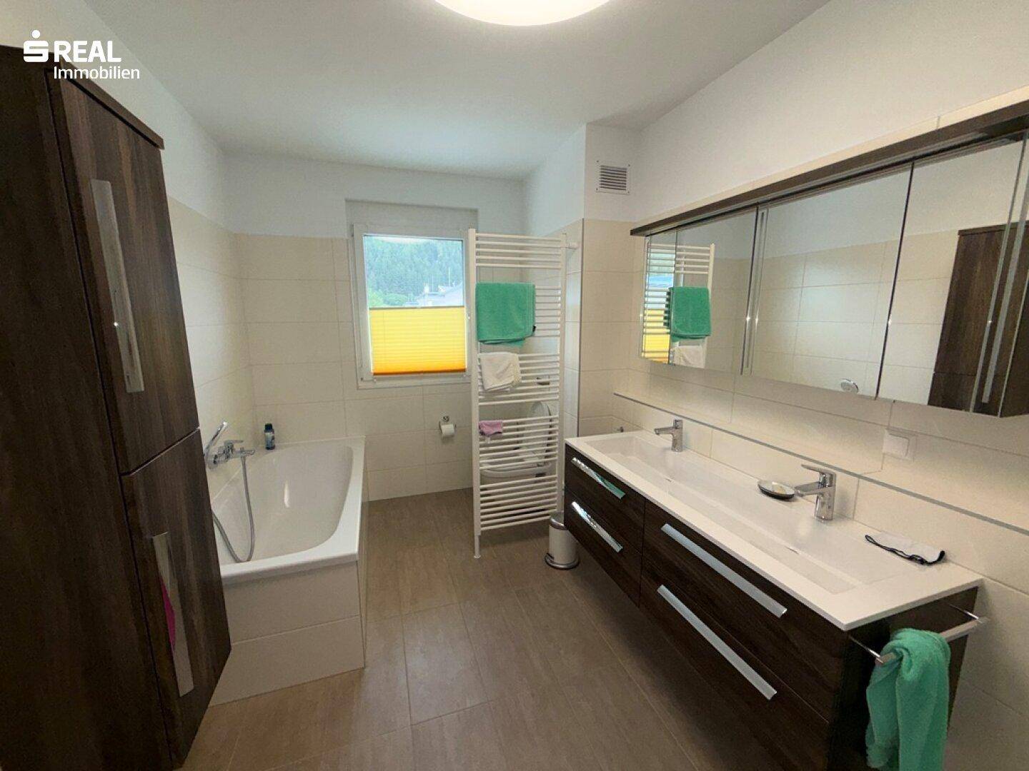 Helles Badezimmer mit Badewanne, Doppelwaschbecken, Fenster und dunklen Holzschränken.