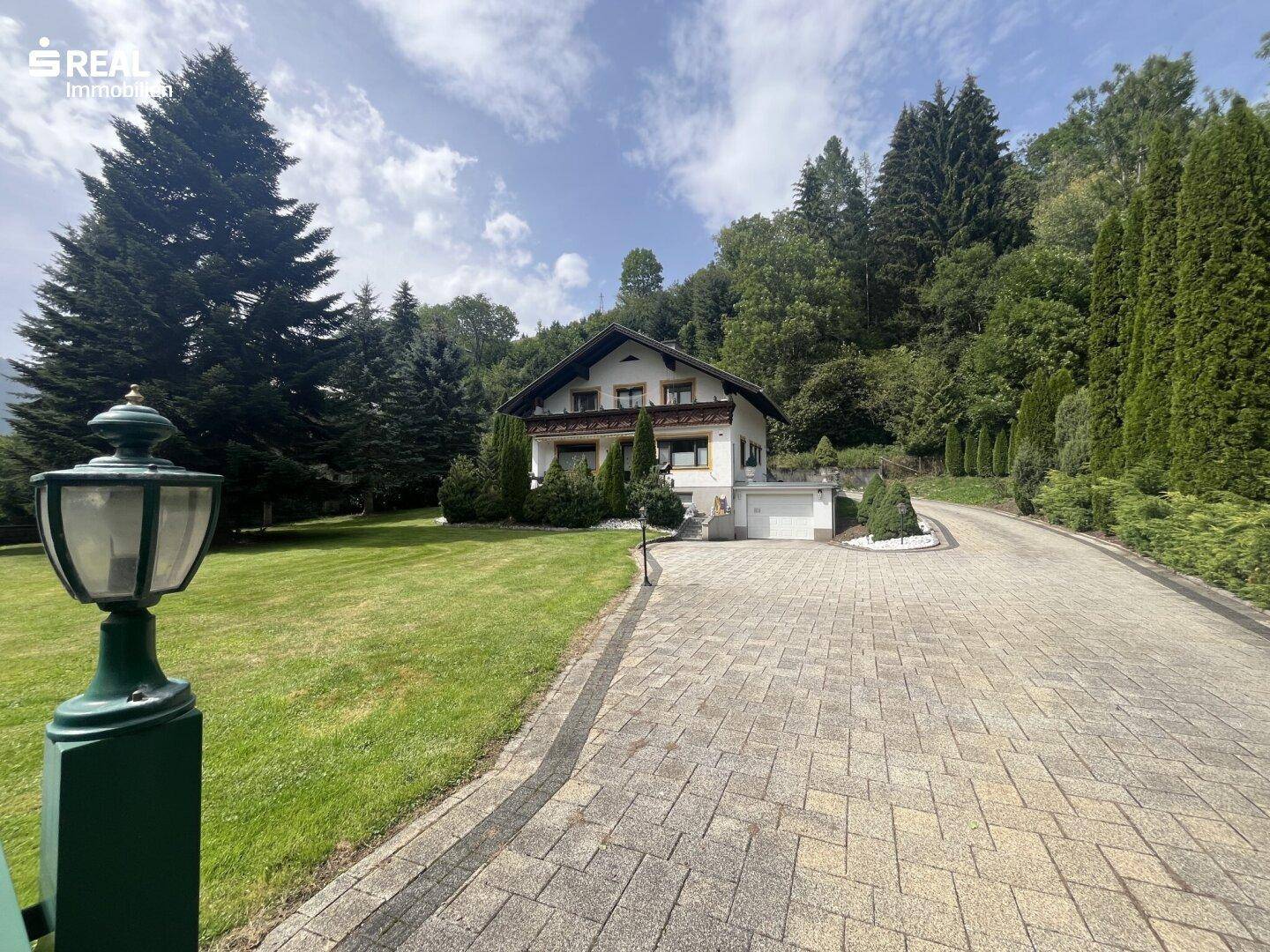 Charmantes Haus mit großer Auffahrt und Laternen, umgeben von einem weitläufigen Garten und Wald.