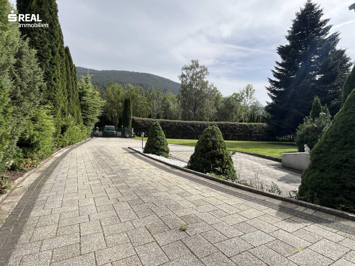 Lange, gepflasterte Auffahrt führt durch einen üppigen Garten mit hohen Hecken und Bäumen.