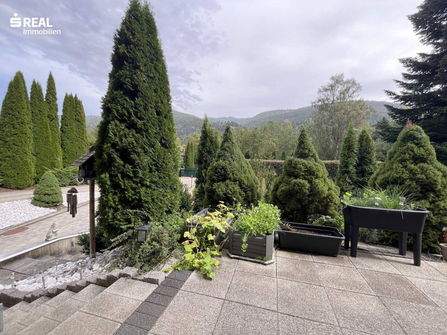 Terrasse mit Kräuterbeeten und Treppenzugang zum Garten, umgeben von hohen Bäumen und Natur.