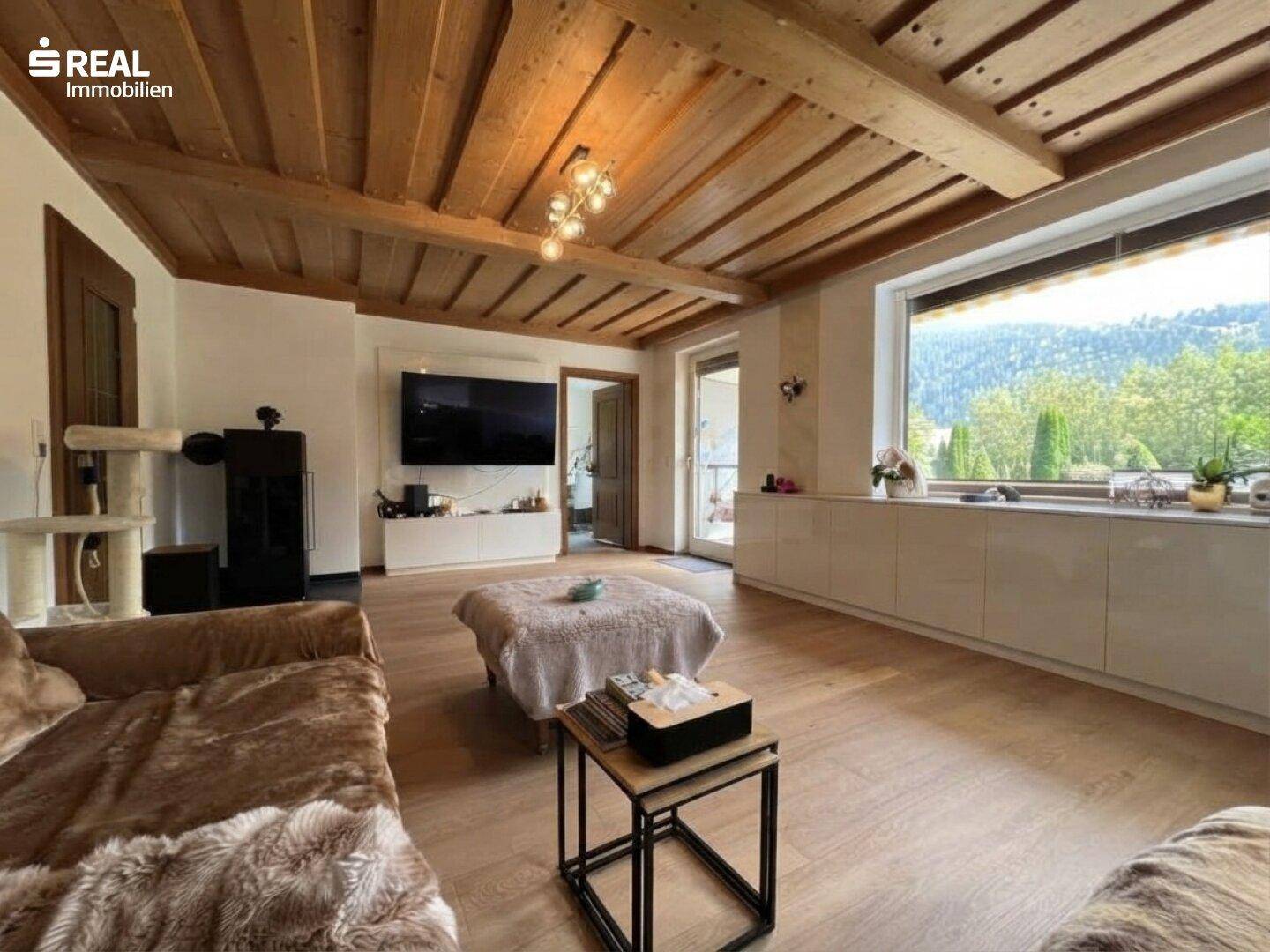 Helles Wohnzimmer mit Holzbalkendecke, großem Fenster und Blick auf die grüne Umgebung.
