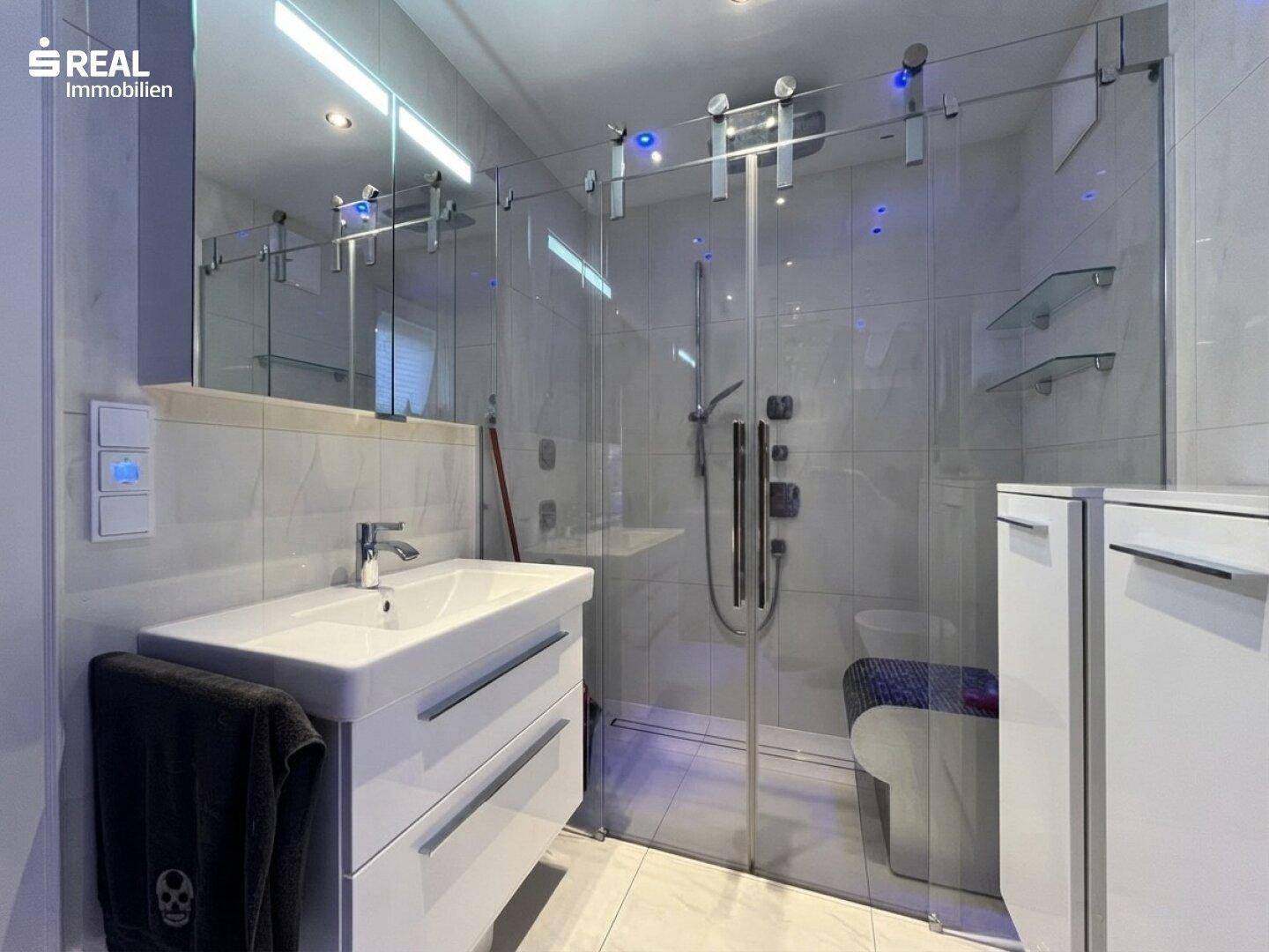 Elegantes Badezimmer mit moderner Glasdusche, Waschtisch und integrierter Beleuchtung.