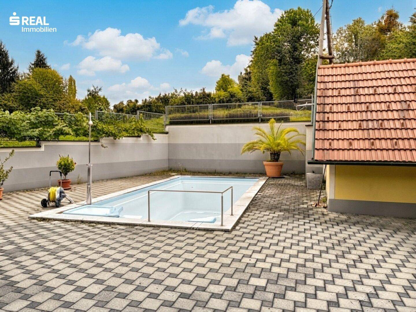 Außenbereich mit eingelassenem Pool, umgeben von einer gepflasterten Fläche und einer kleinen Hütte.
