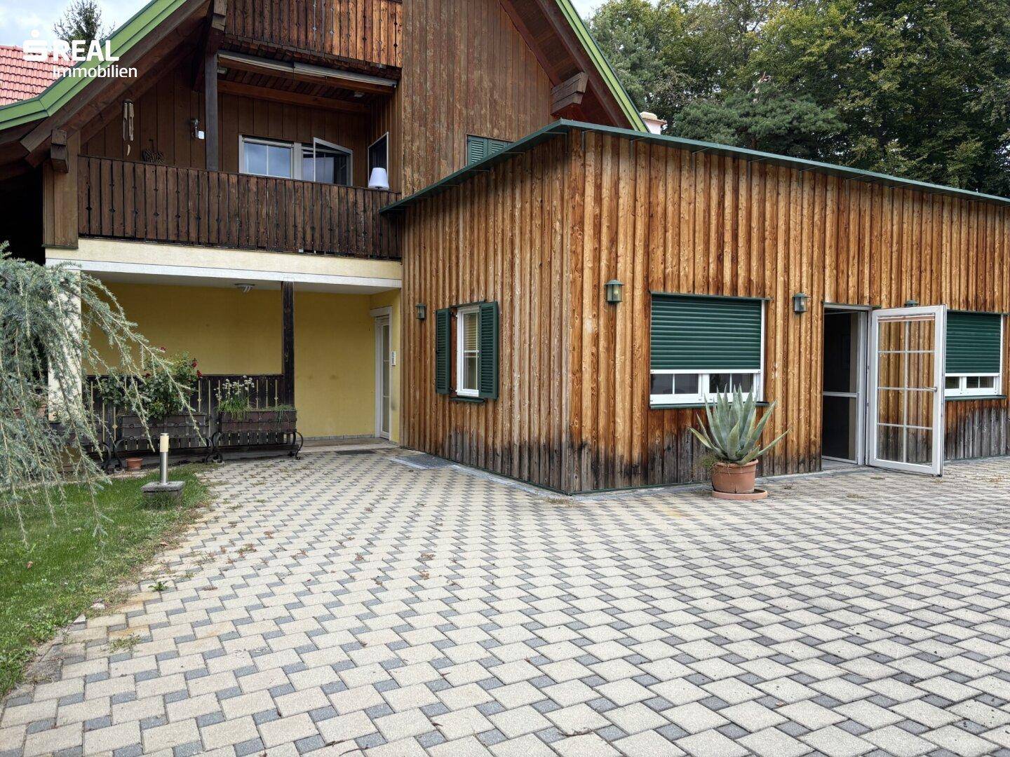 Ansicht des Holzhauses mit gepflastertem Vorplatz und Balkon im Obergeschoss.