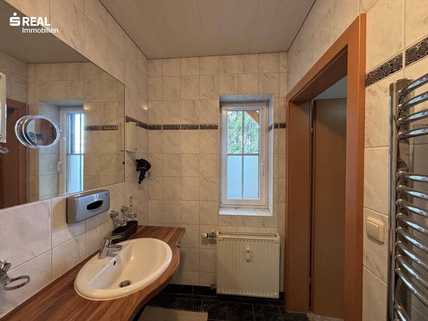 Badezimmer mit Fenster, Waschbecken auf Holzplatte und weißen Fliesen, gut beleuchtet.