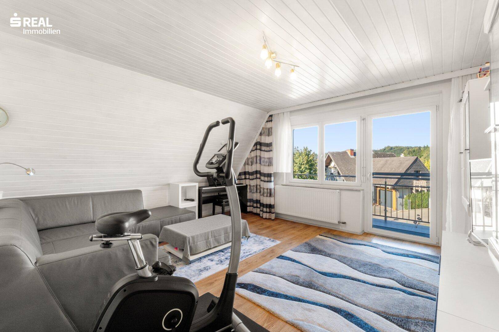 Helles Zimmer mit Sofa, Trainingsgerät und Zugang zum Balkon mit weitem Ausblick.
