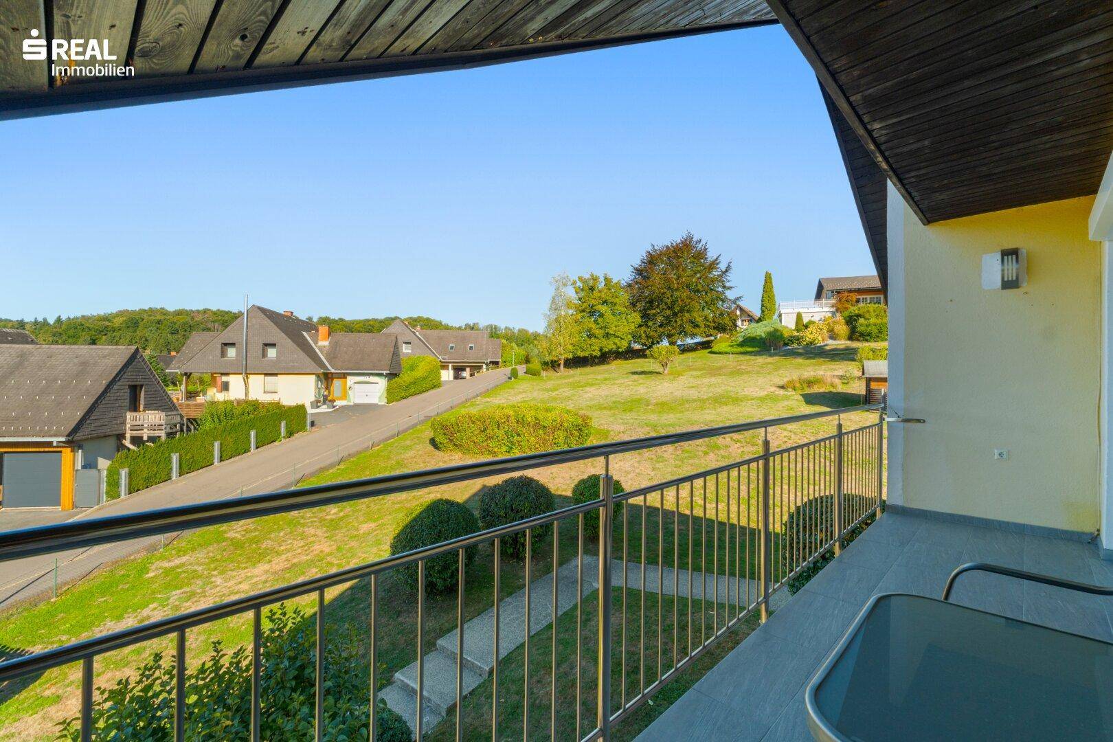 Blick vom Balkon auf die grüne Umgebung und die Nachbarhäuser unter blauem Himmel.