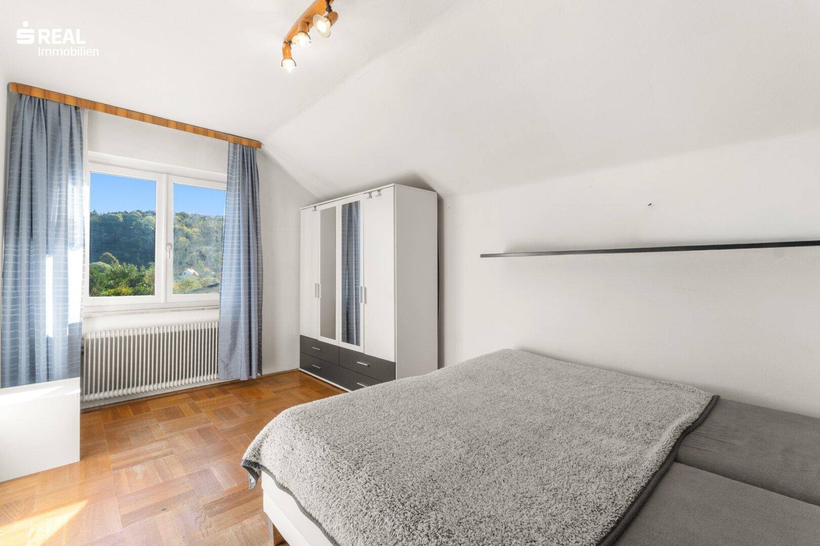 Schlafzimmer mit Parkettboden, Kleiderschrank und Fenster mit Blick ins Grüne.