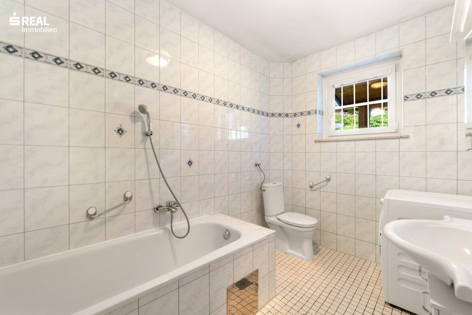 Gefliestes Badezimmer mit Badewanne, Toilette, Fenster und Haltegriffen.