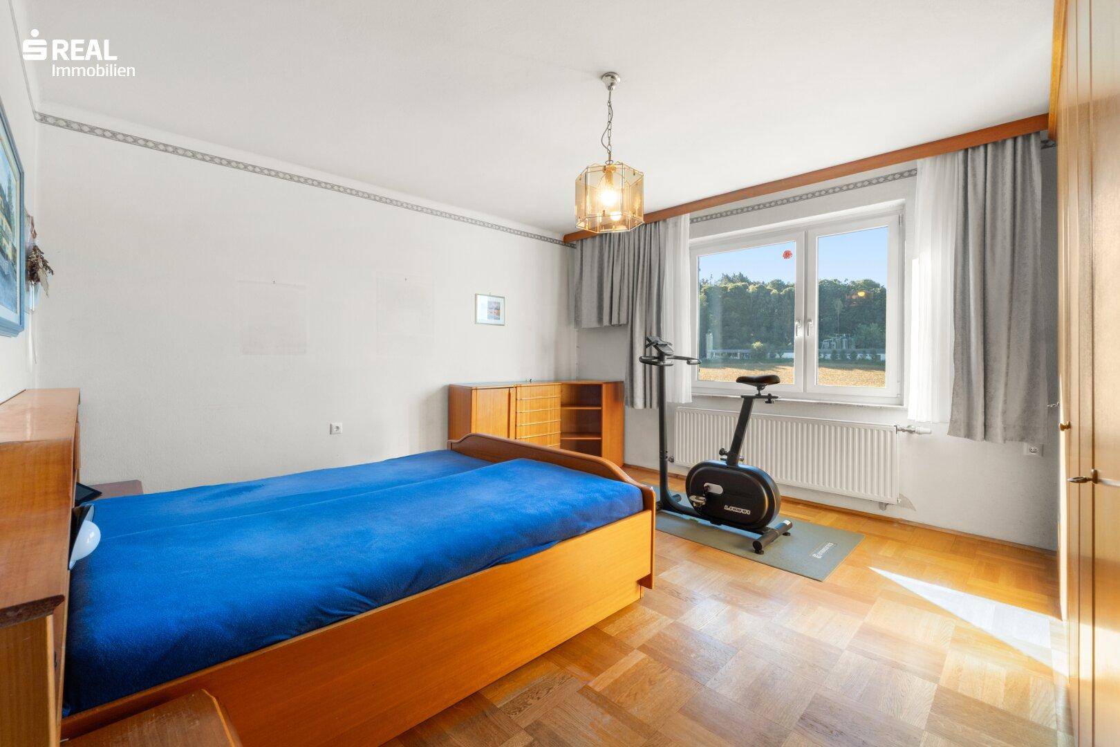Schlafzimmer mit Holzbett, Parkettboden und Fenster mit Blick ins Grüne.