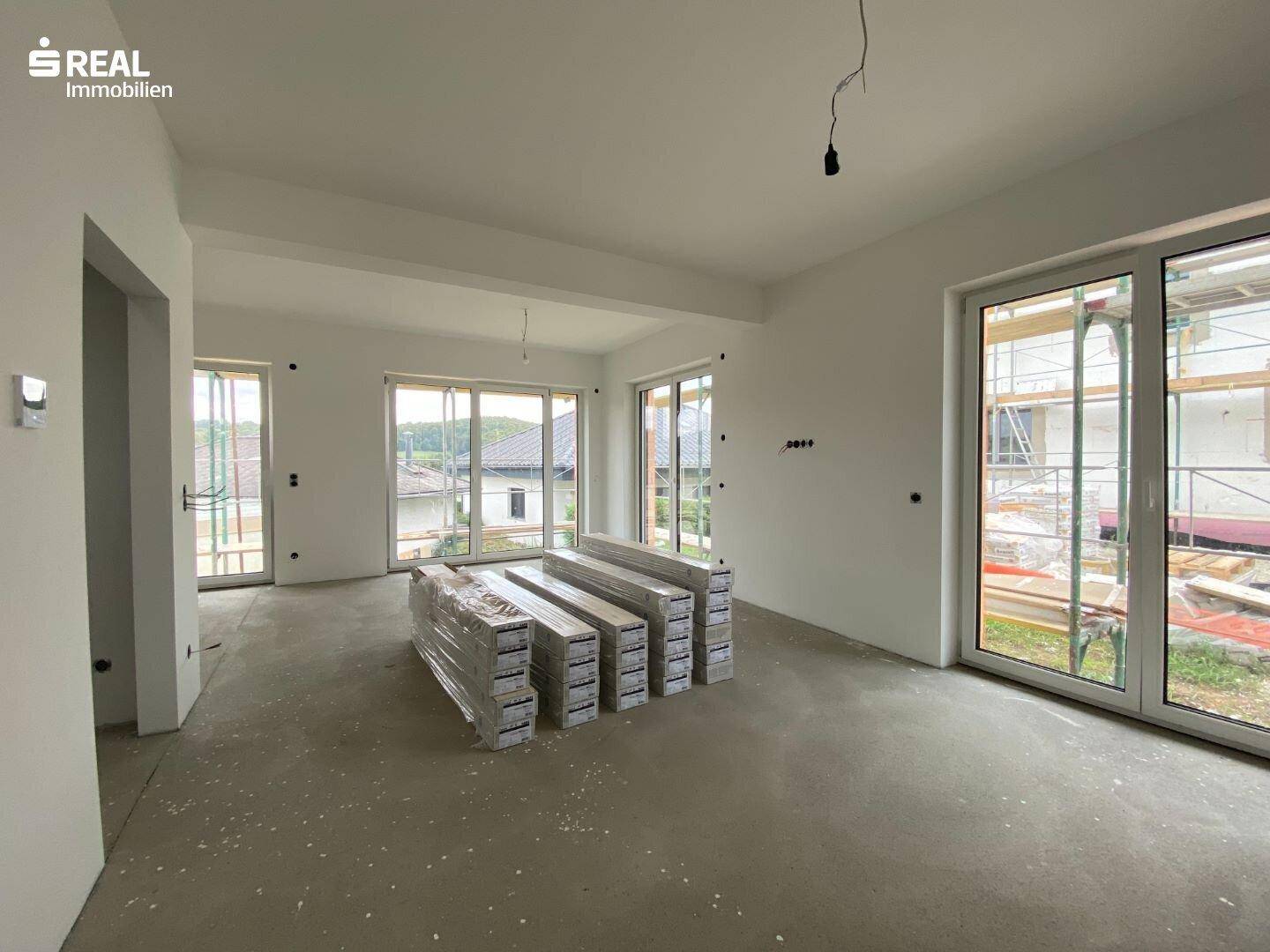 Geräumiger Wohnbereich im Neubau mit großen Fenstern und vorbereitetem Bodenbelag.