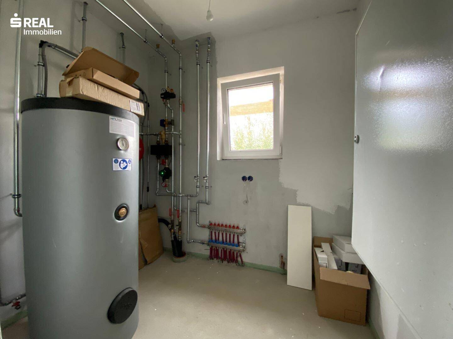 Technikraum mit Warmwasserspeicher, Heizungsinstallationen und einem Fenster.