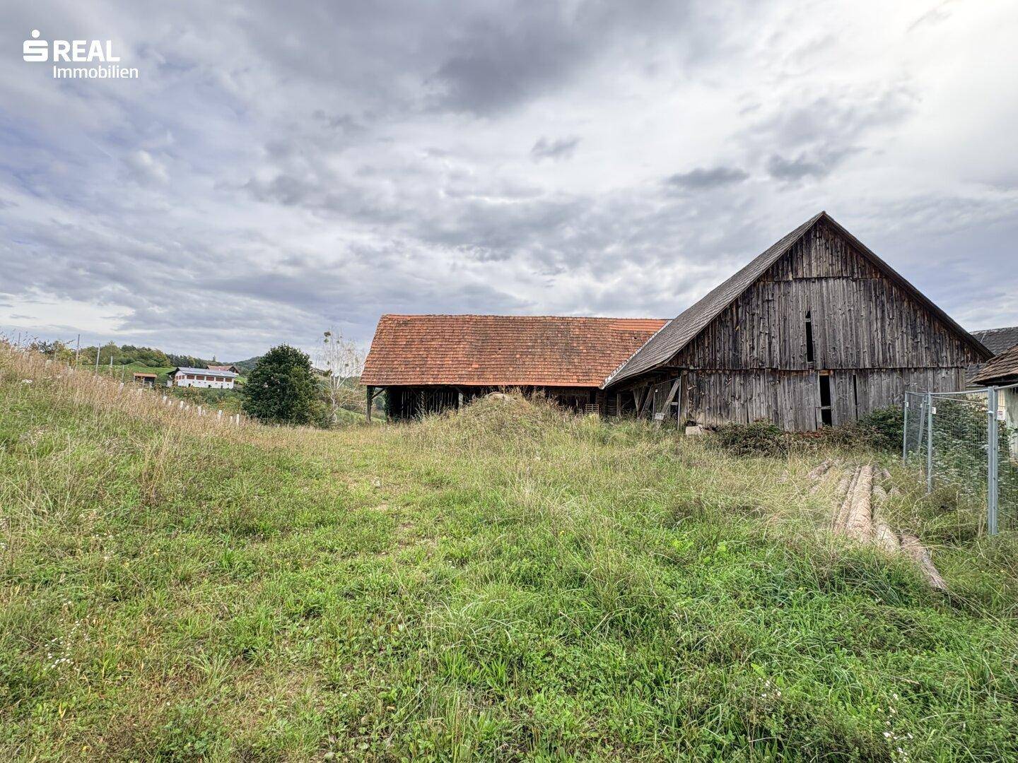 Große Holzscheune und ein weiteres Gebäude mit rotem Ziegeldach auf einem grasbewachsenen Grundstück.