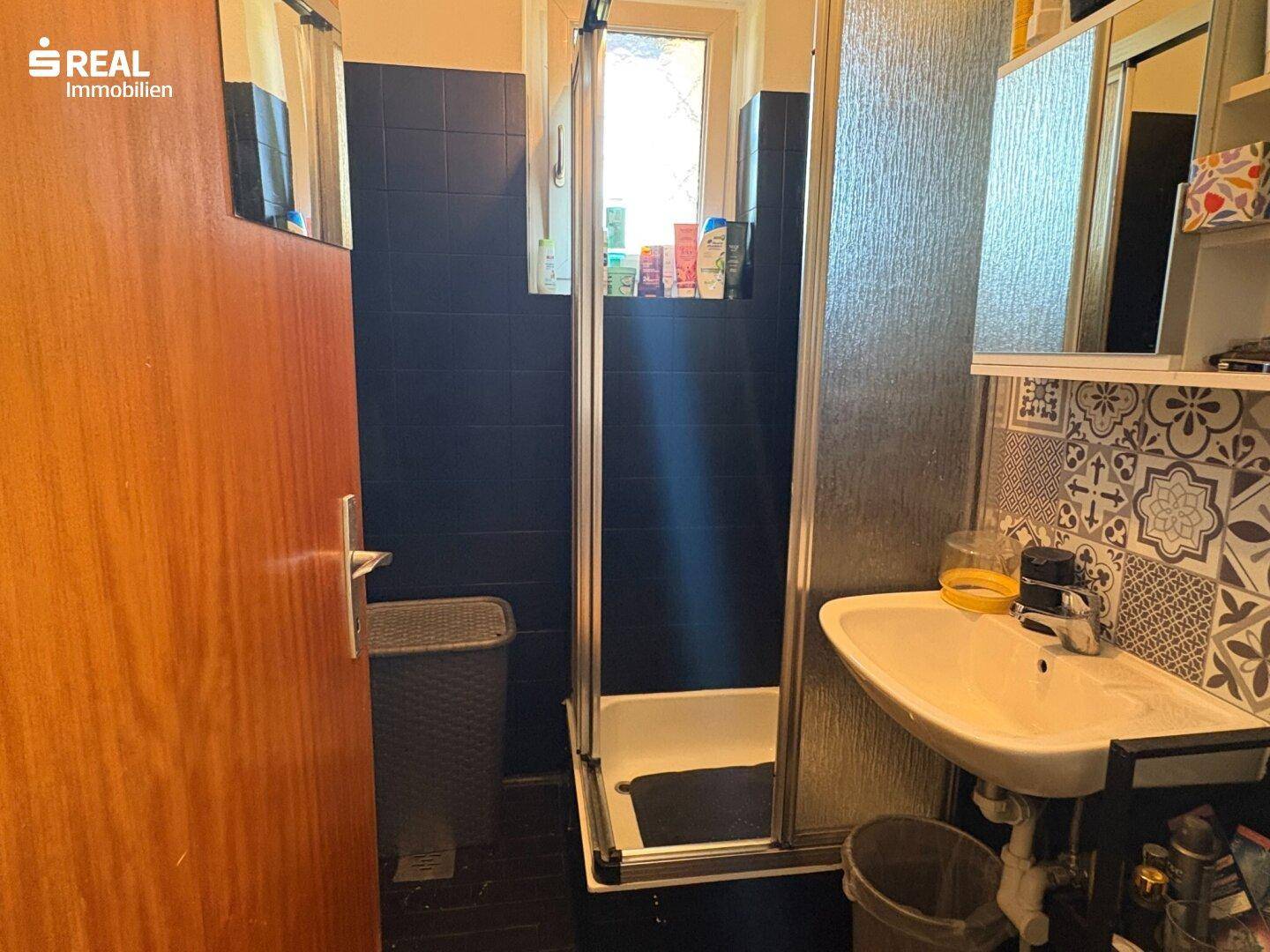 Funktionales Badezimmer mit Dusche, Waschbecken und Fenster, ausgestattet mit dunklen Fliesen.