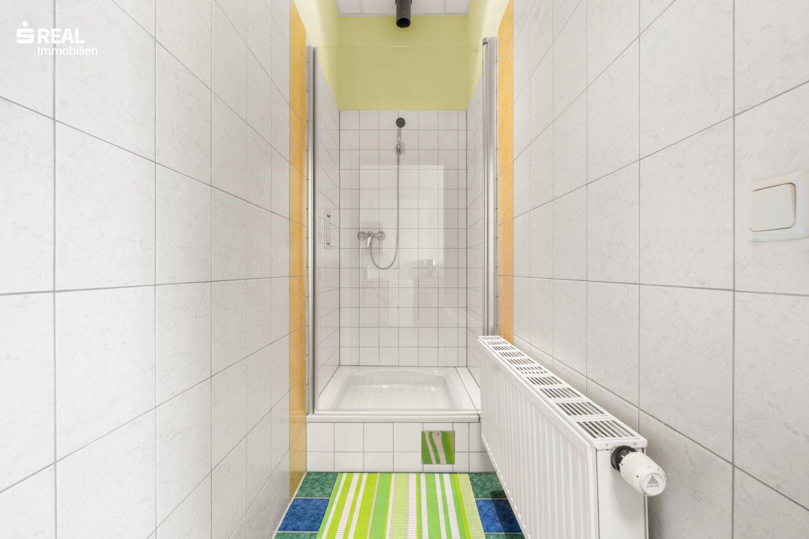 Duschbereich im Badezimmer mit Glasabtrennung, Heizkörper und farbigem Teppich.