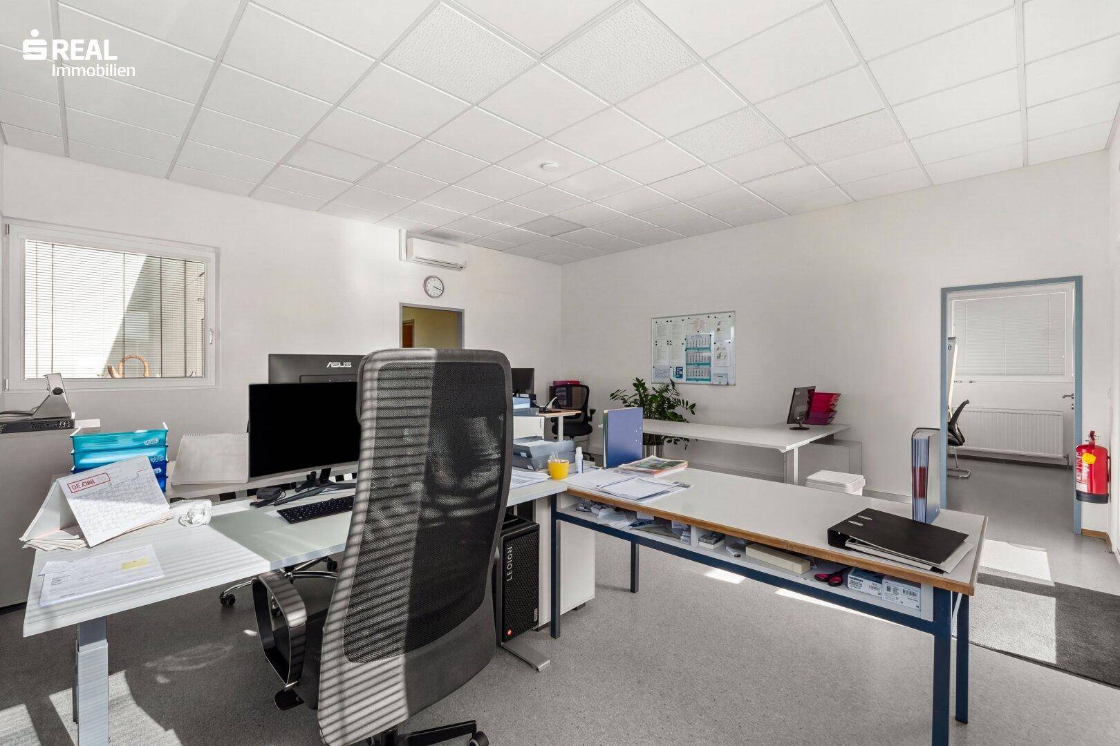 Gut ausgestattetes Büro mit mehreren Schreibtischen, Bürostühlen und einem Fenster mit Jalousien.