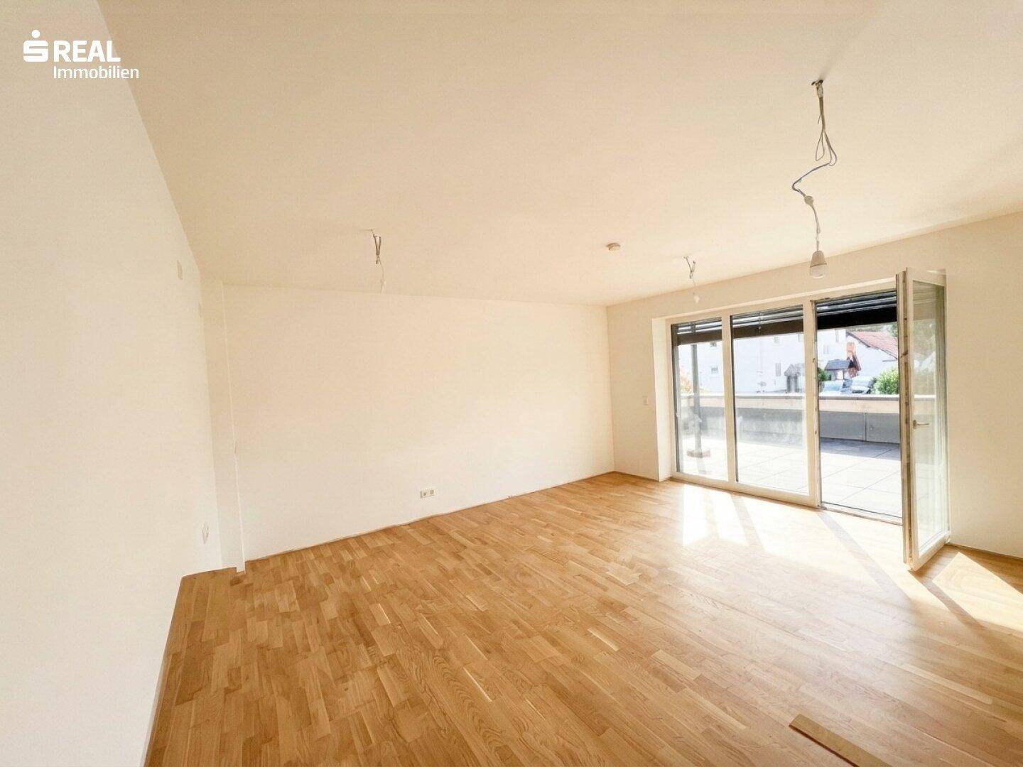 Helles, leeres Wohnzimmer mit großen Fenstern und Zugang zu einer Terrasse, ausgestattet mit Parkettboden.