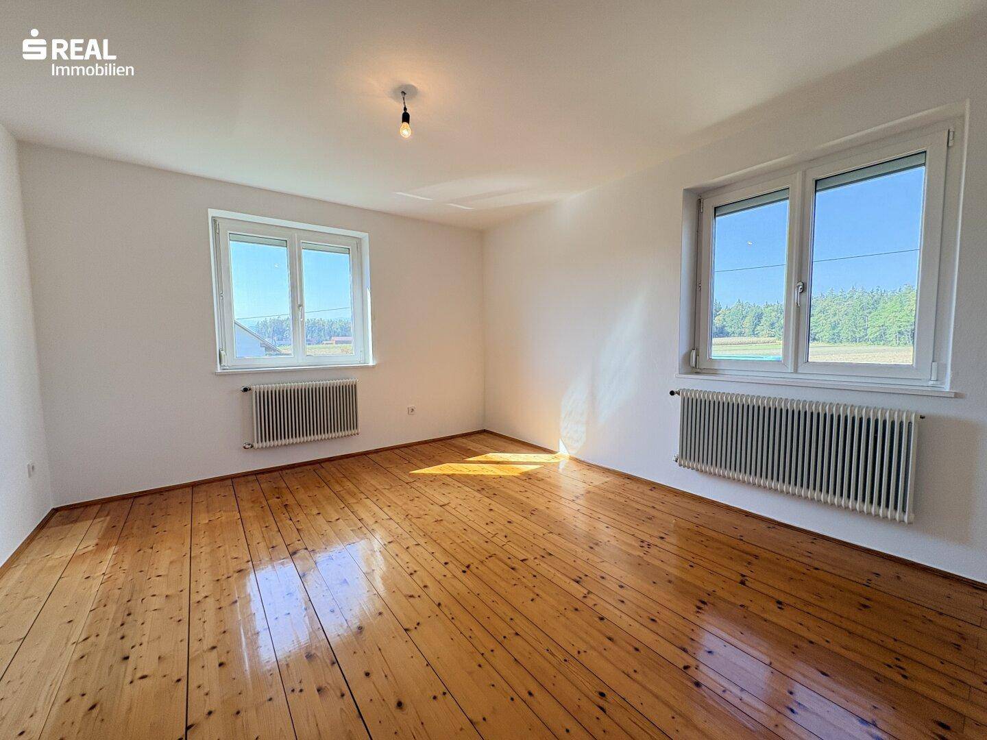 Geräumiges, helles Zimmer mit Holzdielenboden, zwei Fenstern und Blick ins Grüne.