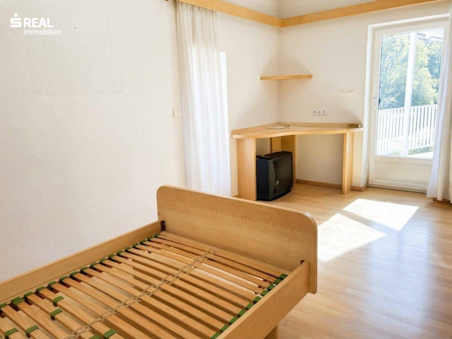 Helles Schlafzimmer mit Holzboden, Schreibtisch und Zugang zum Balkon.