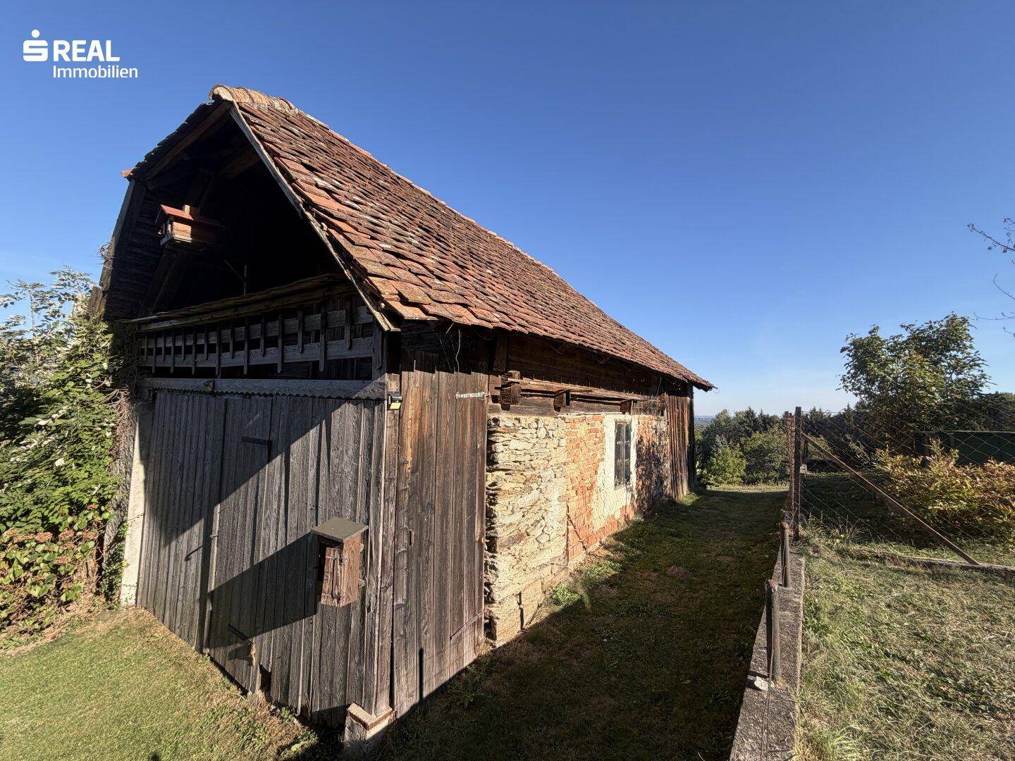 Altes, rustikales Nebengebäude mit Holzwänden und Ziegeldach, umgeben von Gras und Bäumen.