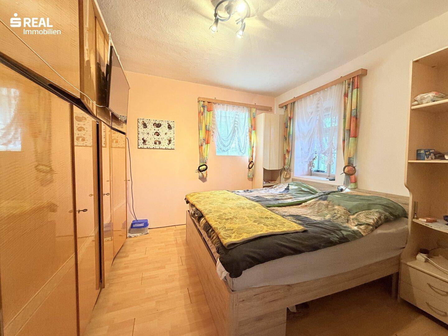 Schlafzimmer mit Doppelbett, Holzschrank und zwei Fenstern, hell und funktional eingerichtet.